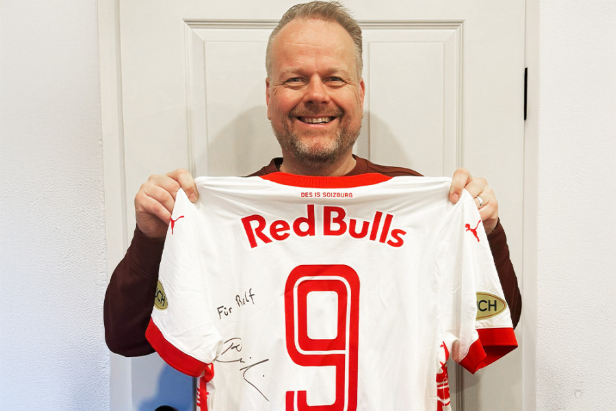 Person hält ein signiertes Red-Bulls-Fußballtrikot mit der Nummer 9 vor einer weißen Tür.