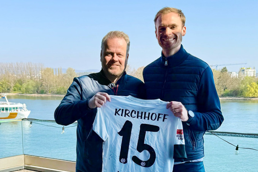 Zwei Personen halten gemeinsam ein weißes Fußballtrikot von Mainz 05 mit der Nummer 15 und dem Namen Kirchhoff vor einer Uferkulisse.   