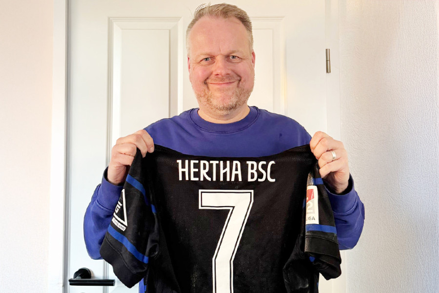 Person hält ein Hertha‑BSC‑Trikot mit der Nummer 7 und dem Namen Niederlechner vor einer weißen Tür.
