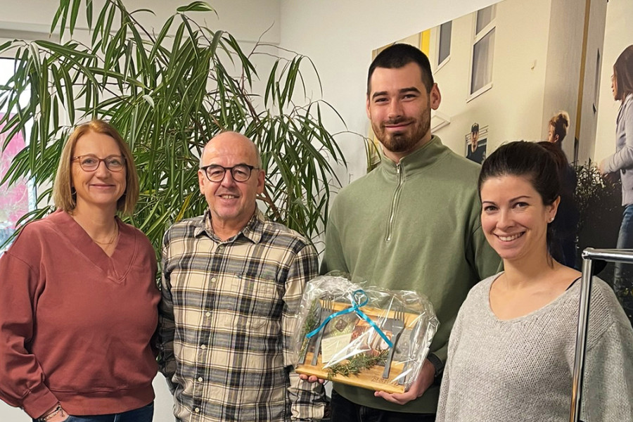 Gruppenfoto im Büro mit Geschenkübergabe, dekoriertem Präsentkorb und Pflanzen im Hintergrund.