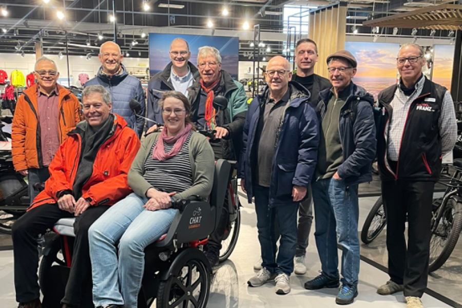 Gruppe von Personen steht in einem Fahrradgeschäft um eine moderne Rollstuhl-Rikscha, in der zwei Personen sitzen.
