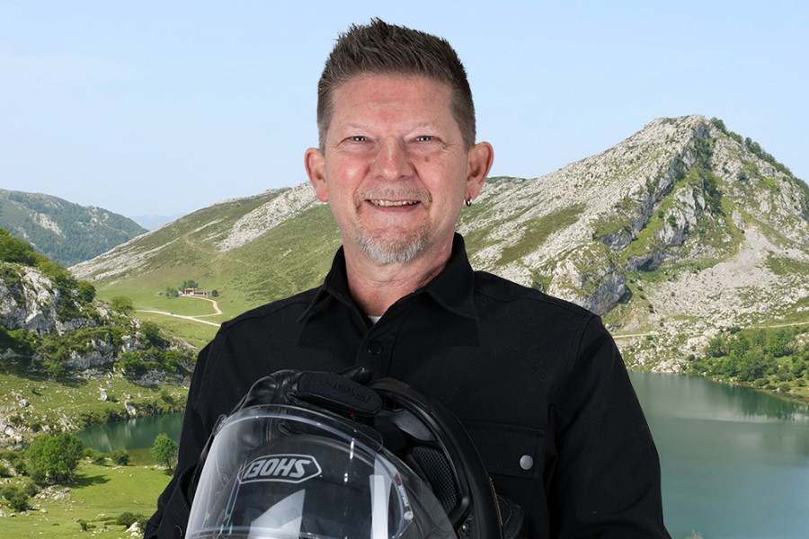 Person hält einen Motorradhelm und steht vor einer Berglandschaft mit See und Felsformationen.