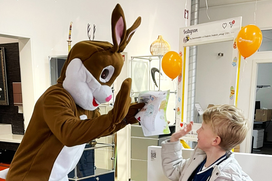 Osterhase im Kostüm gibt einem Kind ein High‑Five und begutachtet ein gemaltes Osterbild in einem Geschäft.