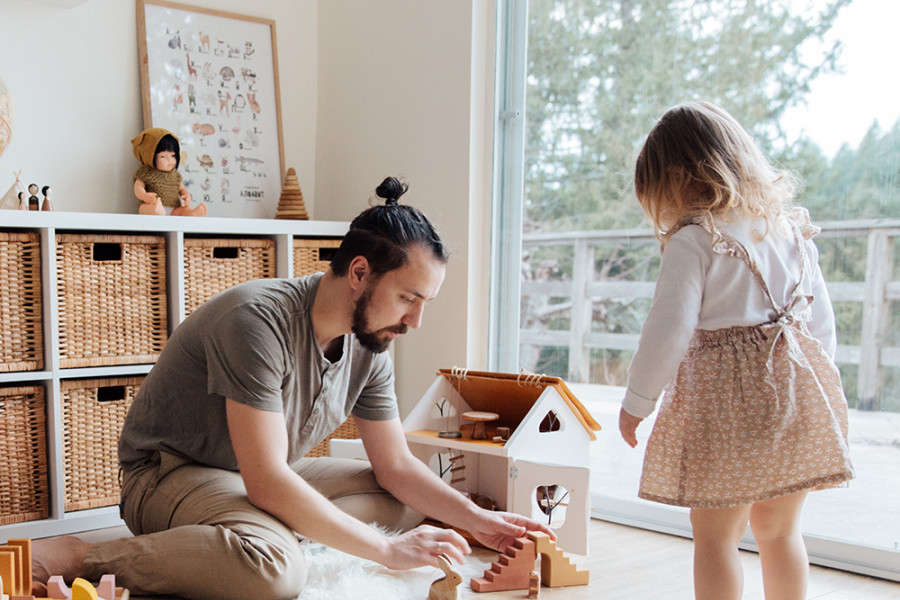 Zwei Personen spielen in einem hellen Wohnzimmer mit einem Holzspielhaus und Bauklötzen auf dem Boden.