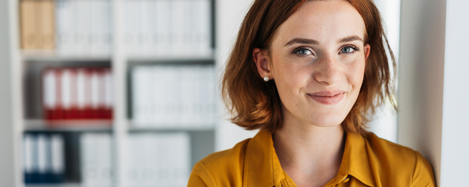 Frau in gelber Bluse steht in einem modernen Büro vor einem Regal mit Aktenordnern.