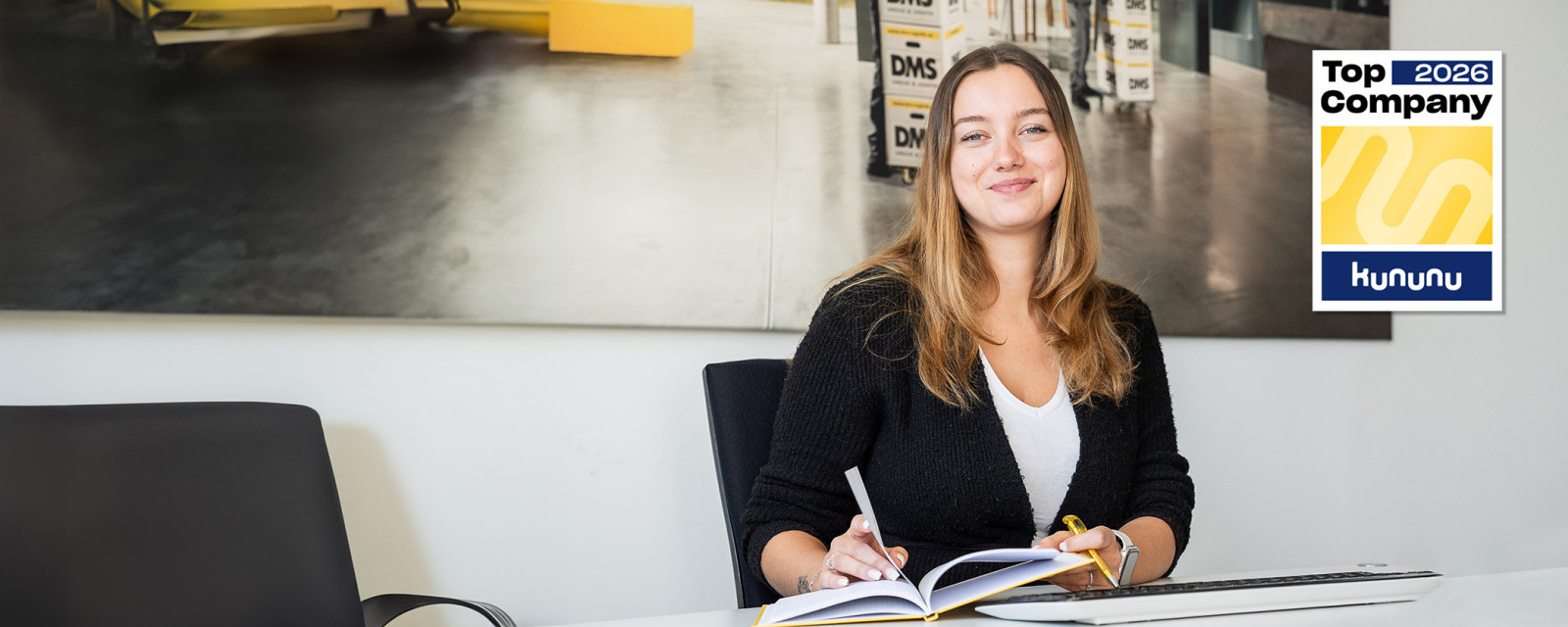 Mitarbeiterin arbeitet am Schreibtisch mit Notizbuch und Tastatur, modernes Büro mit Logistik‑Motiv im Hintergrund und Kununu‑Top‑Company‑2026‑Siegel.